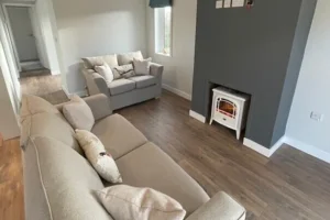 Living room with beige sofas, a dark accent wall with a small fireplace, and hardwood floors, illuminated by natural light from a window.