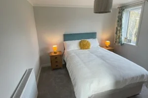 Double bed with white bedding, blue headboard, two bedside tables with yellow lamps, window with floral curtains, and a ceiling light fixture.