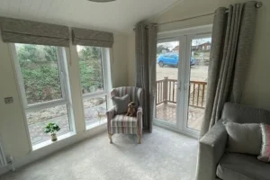A cozy living room corner with large windows, a striped armchair with a small pillow and stuffed dog, gray sofa, and a wooden deck outside.