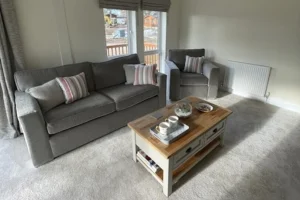 A living room with a gray sofa, an armchair, a wooden coffee table, and large windows with curtains, sunlight, and outdoor view.