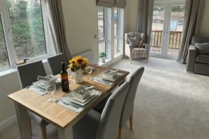 Dining table set with plates, glasses, a bottle of wine, and a sunflower centerpiece in a bright living room with large windows and a balcony.