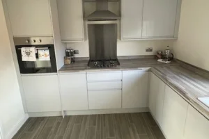 Modern L-shaped kitchen with white cabinets, stainless steel range hood, built-in oven, and wooden countertop, set against light-colored walls and flooring.