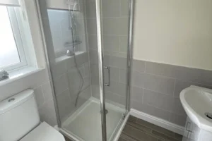 A small bathroom with a glass shower enclosure, a white toilet, a wall-mounted sink, a frosted window, and gray tiled walls and floor.