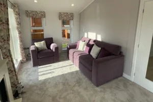 A cozy living room with two purple sofas, light-colored walls, floral curtains, and sunlight streaming through windows, creating a warm and inviting atmosphere.