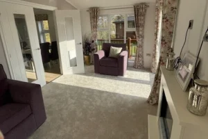 A cozy living room with purple armchairs, floral curtains, a beige carpet, and a white fireplace mantel decorated with framed photos and candles.