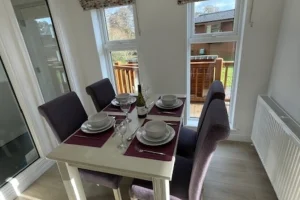 Dining table set for four with plates, glasses, wine bottle, and cutlery in a bright room with large windows and a sliding door leading to an outdoor deck.