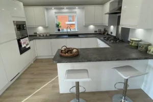 Modern white kitchen with a dark countertop island, two white bar stools, and a window with a small plant in the background.