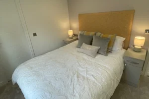 A neatly made bed with a fabric headboard, multiple pillows, and two bedside tables with lamps in a cozy, softly lit bedroom.
