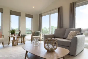 A bright living room with large windows, beige curtains, a gray sofa with throw pillows, a wooden coffee table with decorative items, and outdoor scenery visible through the windows.