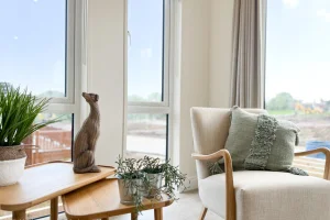 A cozy living room corner with a beige armchair, green pillows, a wooden side table with potted plants and a decorative dog sculpture, near large windows with curtains.