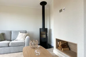 A cozy living room with a grey sofa, wooden coffee table, a wood-burning stove, and a small storage nook with logs, all under neutral-colored walls.