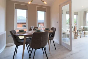 Dining area with a wooden table set with bowls and glasses, four upholstered chairs, large windows, and an adjoining bright living room.