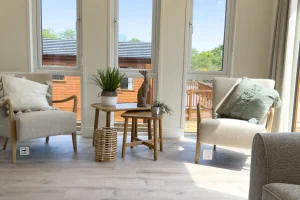 Bright living room with three large windows, two armchairs with cushions, and a small round table with plants and decor, sunlight streaming in.