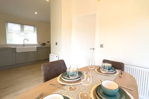 A dining table set for four with bowls, plates, wine glasses, and cutlery, in a bright, modern kitchen and dining area with neutral tones and minimalist decor.