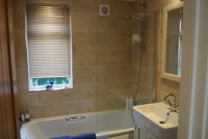 Bathroom with beige tiled walls, a window with closed blinds, a small bathtub, and a white sink with a mirror above.