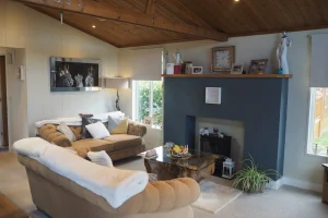 Cozy living room with beige sofas, a dark blue fireplace, wooden ceiling, and shelves with pictures and decor, lit by natural light from windows.