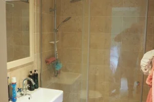 Bathroom with a corner shower enclosure, a white sink with toiletries, and a mirror. Beige tiled walls and ceiling, with a partial view of a towel hanging on the right.