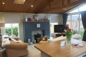 A cozy living room with a wooden vaulted ceiling, beige sofa, TV, fireplace, and a table with a vase of red and white tulips, large windows, and garden view.