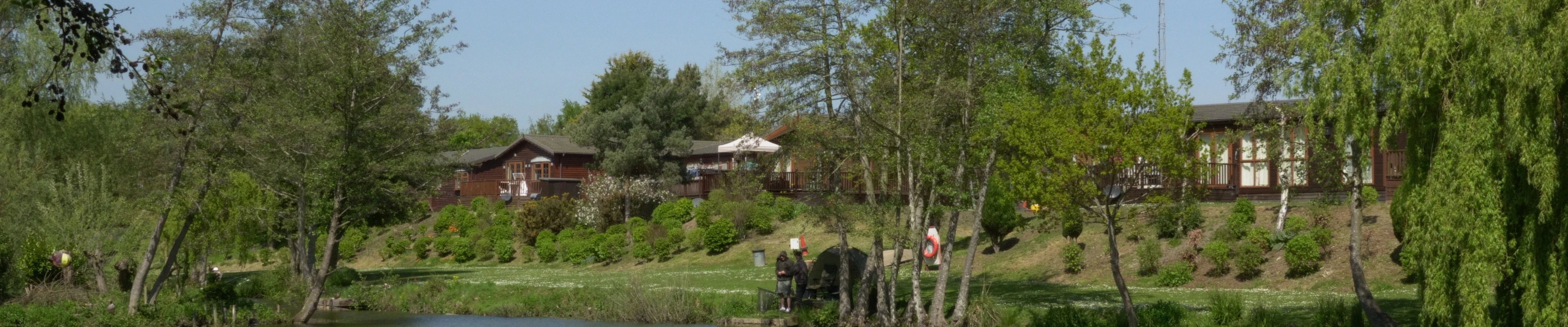 A peaceful lakeside scene with trees, houses, and people enjoying the outdoors under a clear blue sky.