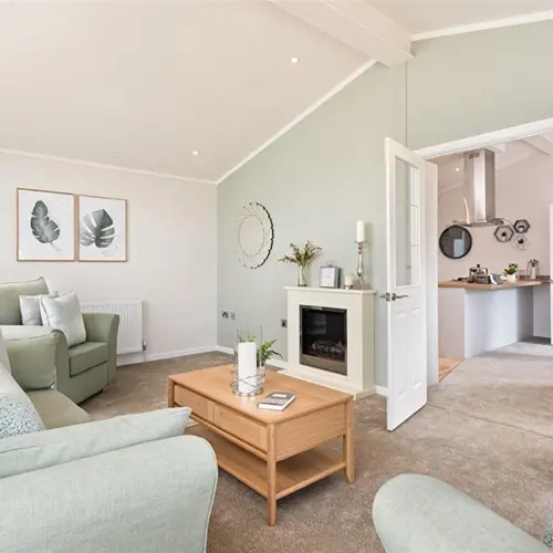 Living room with light-colored sofas, artwork of leaves on the wall, a wooden coffee table, and open double doors leading to a kitchen with modern decor.