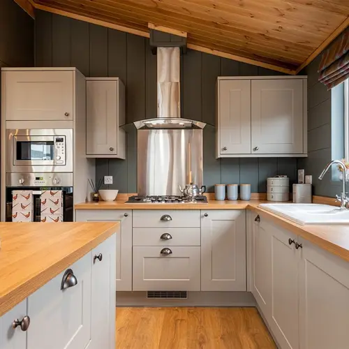 A cozy kitchen with white cabinets, a wooden countertop, stainless steel appliances, a window with striped curtains, and a sloped wooden ceiling.