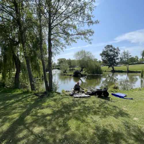 A person is sitting on a chair near a calm lake surrounded by trees, with fishing gear and a blue flotation device on the grass in a sunny park.