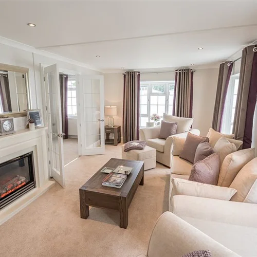 Bright living room with cream sofas, colorful cushions, large windows with curtains, a fireplace, and a wooden coffee table.