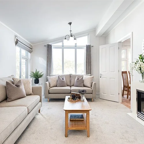 Bright living room with beige sofas, a wooden coffee table, large windows, and elegant decor including a round mirror and floral arrangement.