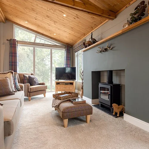 Living room with a high, angled wooden ceiling, a fireplace with decorative items on the mantel, a large window with colorful curtains, and cozy seating around a TV.