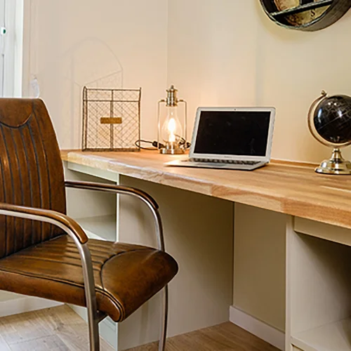 A cozy workspace with a wooden desk, brown leather chair, laptop, globe, lamp, and decorative items in a well-lit room.