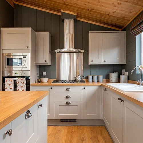 A cozy kitchen with white cabinets, a wooden countertop, stainless steel appliances, a window with striped curtains, and a sloped wooden ceiling.