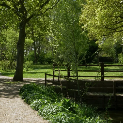 A peaceful park scene with a gravel pathway, lush green trees, a small wooden bridge over a waterway, and sunlight filtering through the foliage.