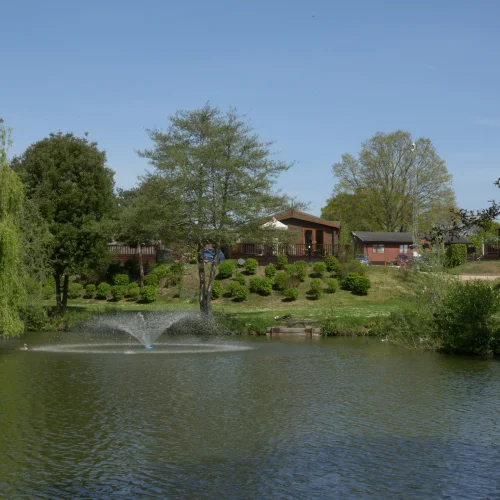 A peaceful lakeside scene with trees, a fountain in the water, and houses on a hill in the background under a clear blue sky.