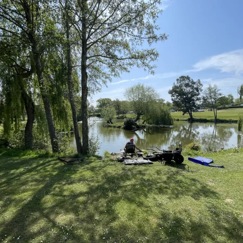 A person is sitting on a chair near a calm lake surrounded by trees, with fishing gear and a blue flotation device on the grass in a sunny park.