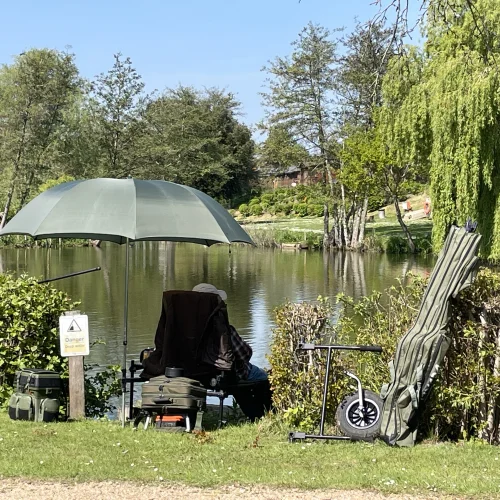 A person is sitting by a river with an umbrella, surrounded by trees and bushes, with fishing equipment and a sign that warns of danger near the water.