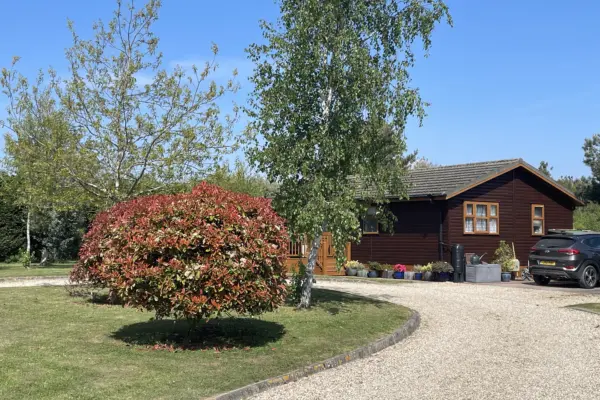 A gravel driveway curves past a vibrant red and green shrub, with a wooden house, parked car, and potted plants in the background under a blue sky.