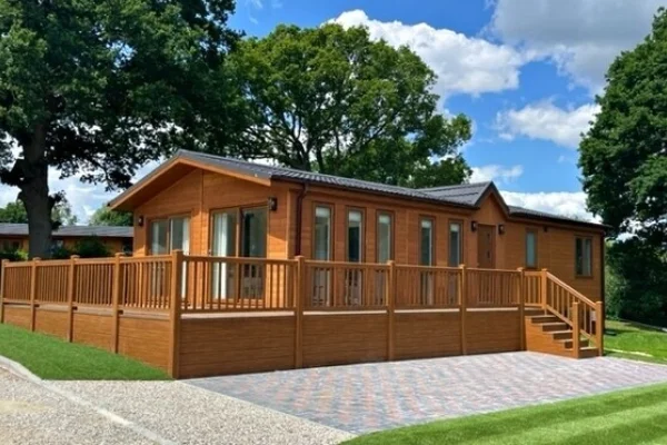 A wooden house with a spacious raised deck, set in a lush green yard with tall trees and a bright blue sky with clouds.