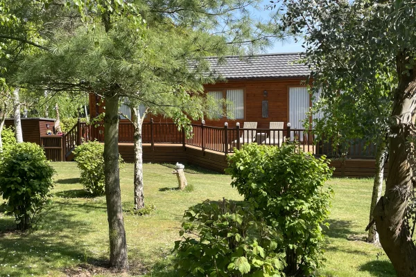 A backyard with a wooden house, a deck with patio furniture, surrounded by trees, bushes, and a grassy lawn on a sunny day.