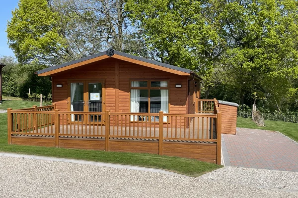 A wooden cabin with large glass doors and windows, surrounded by a wooden fence, set in a lush green area with tall trees and a clear blue sky.