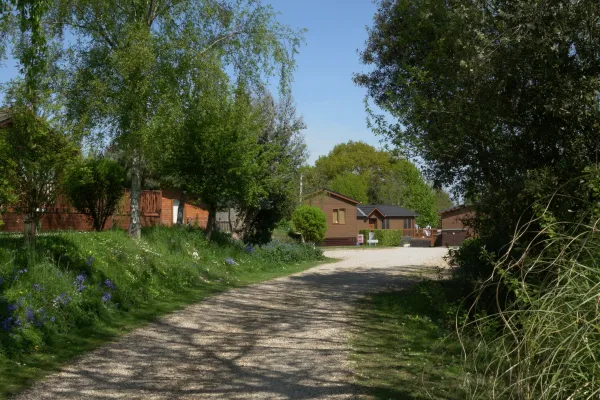 A gravel pathway curves through lush green trees and flowering plants toward small wooden houses under a clear blue sky.