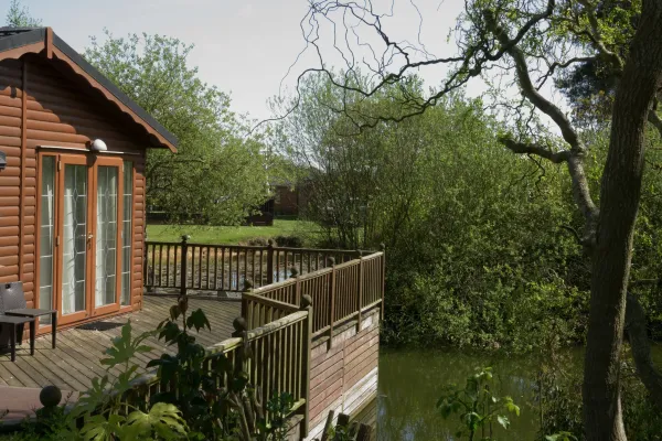 A wooden deck with a railing extends over a pond next to a house with brown logs and glass double doors, surrounded by lush green trees and foliage.