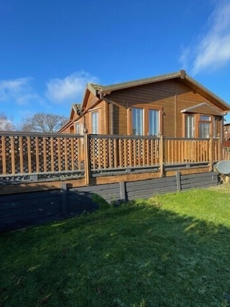 A wooden house with a deck, large windows, a satellite dish, and a grassy yard under a partly cloudy blue sky.