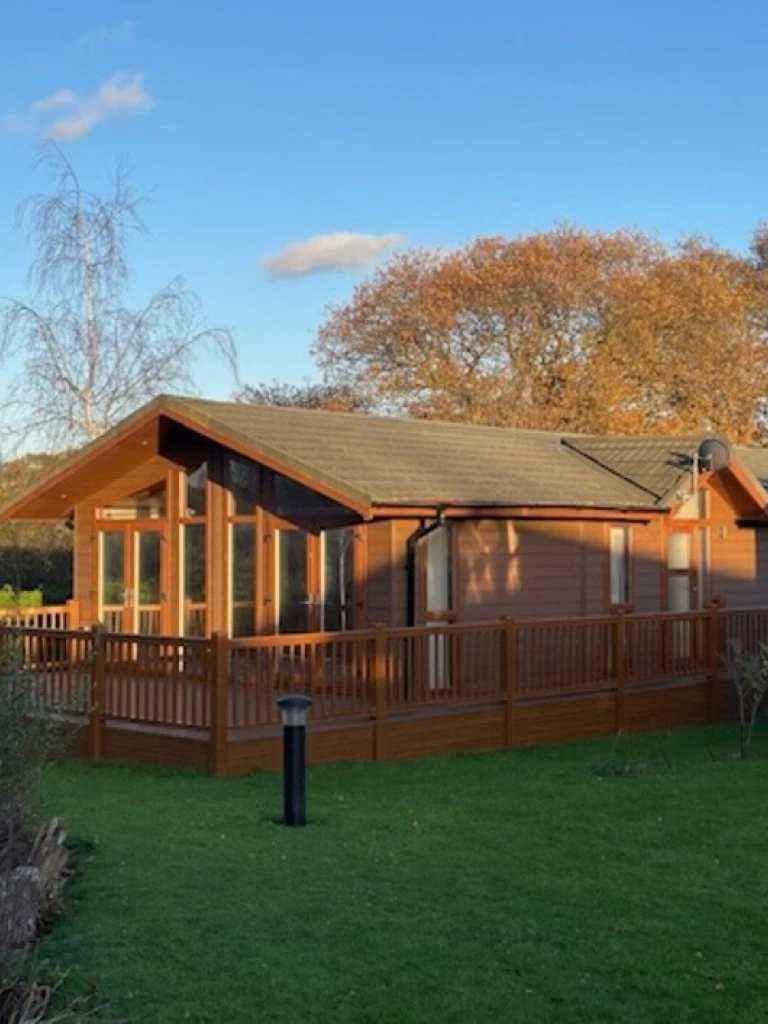 A wooden house with large windows and a deck, surrounded by green grass and trees with autumn leaves, under a clear blue sky.