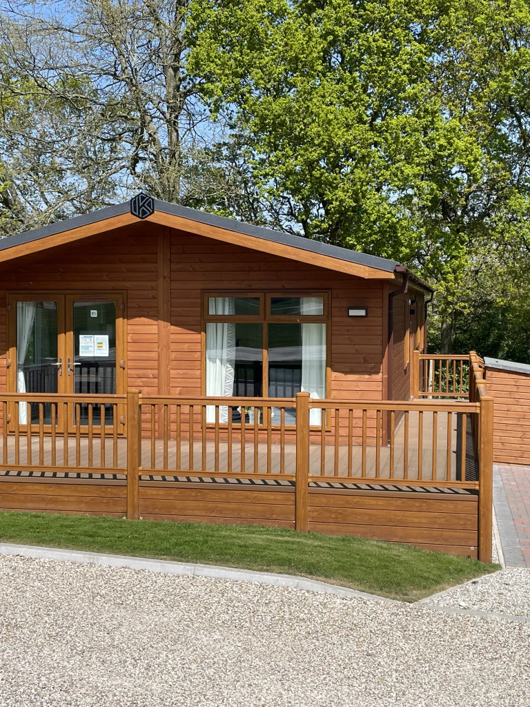 A wooden cabin with large glass doors and windows, surrounded by a wooden fence, set in a lush green area with tall trees and a clear blue sky.