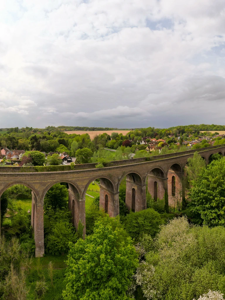 A long stone viaduct arching over a lush green rural landscape with a village and countryside under a cloudy sky.