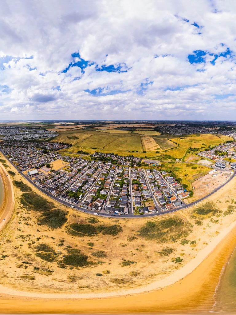 Aerial view of a coastal town with houses, sandy beaches, breaking waves, clear sky, and scattered clouds on a sunny day.